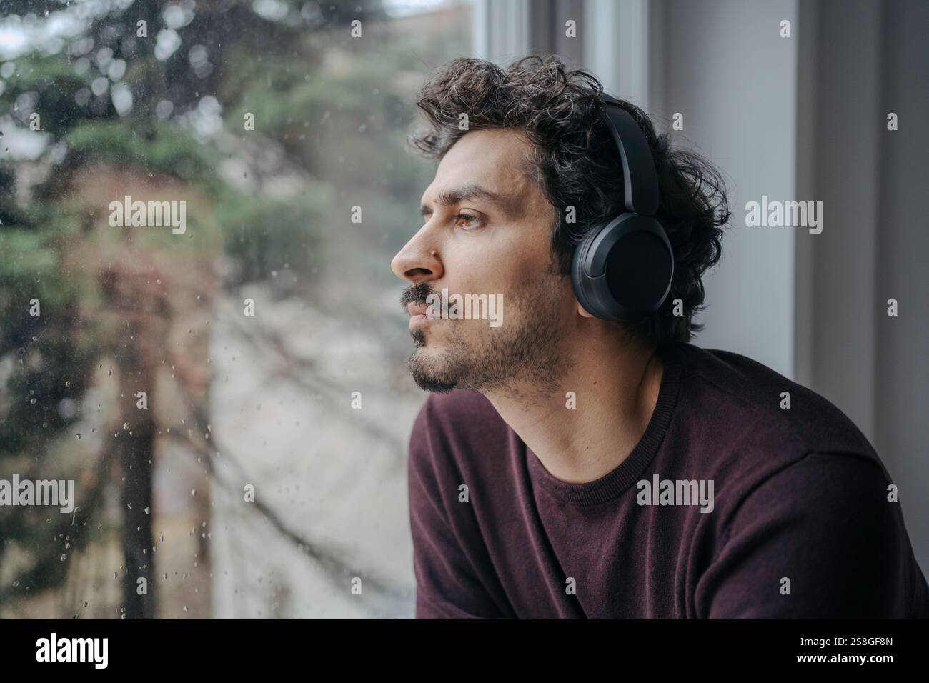 Bell'uomo mediorientale con cuffie sovrauricolari che ascoltano musica e si rilassano vicino alla finestra. Foto Stock