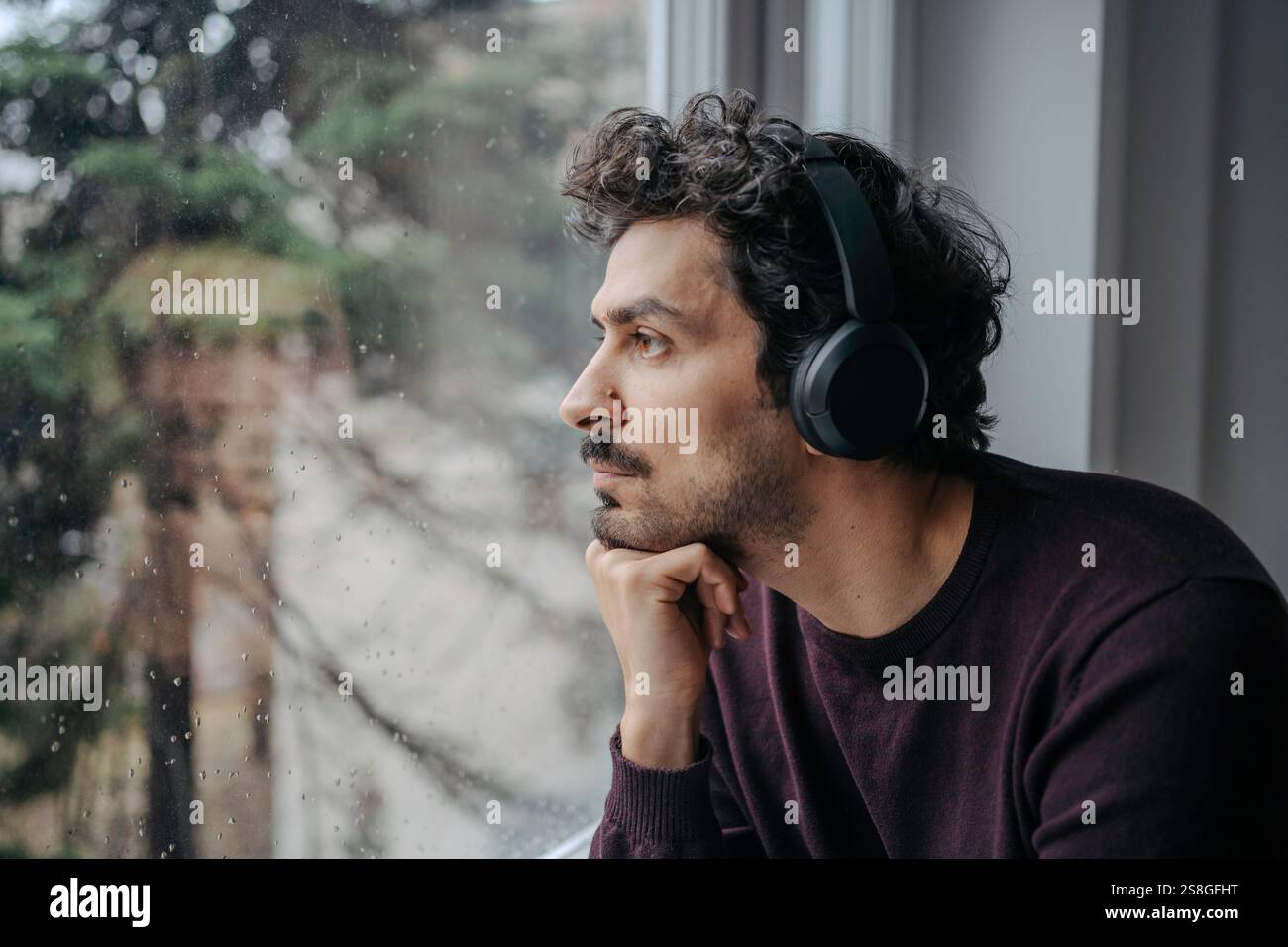 Bell'uomo mediorientale con cuffie sovrauricolari che ascoltano musica e si rilassano vicino alla finestra. Foto Stock