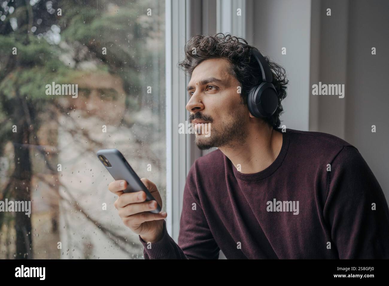 Bell'uomo mediorientale con cuffie sovrauricolari che ascoltano musica e si rilassano vicino alla finestra. Foto Stock