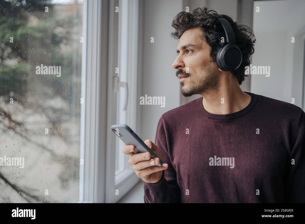 Bell'uomo mediorientale con cuffie sovrauricolari che ascoltano musica e si rilassano vicino alla finestra. Foto Stock