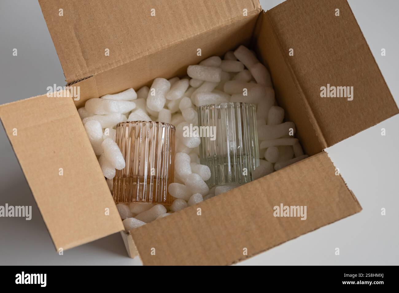 Vetreria fragile in tonalità pastello accuratamente confezionata con arachidi di schiuma protettiva all'interno di una scatola di cartone aperta. Preparazione per la spedizione di articoli delicati Foto Stock