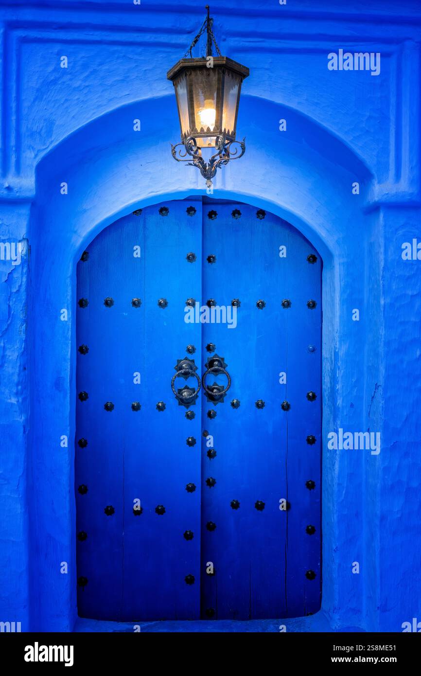 Vista sulla strada di una splendida porta d'ingresso blu in legno illuminata da una vecchia lampada. Architettura e colori blu della Medina, Chefchaouen, Marocco. Casa marocchina. Foto Stock