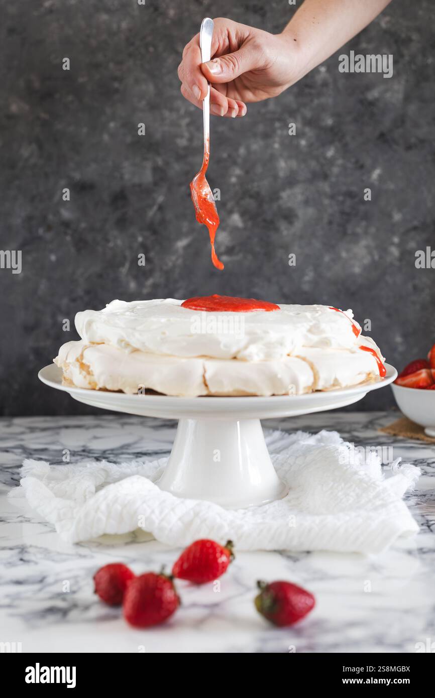 Una mano che aggiunge con cura la salsa di fragole su una pavlova ricoperta di panna montata, adagiata su un piedistallo bianco su una superficie di marmo. Foto Stock