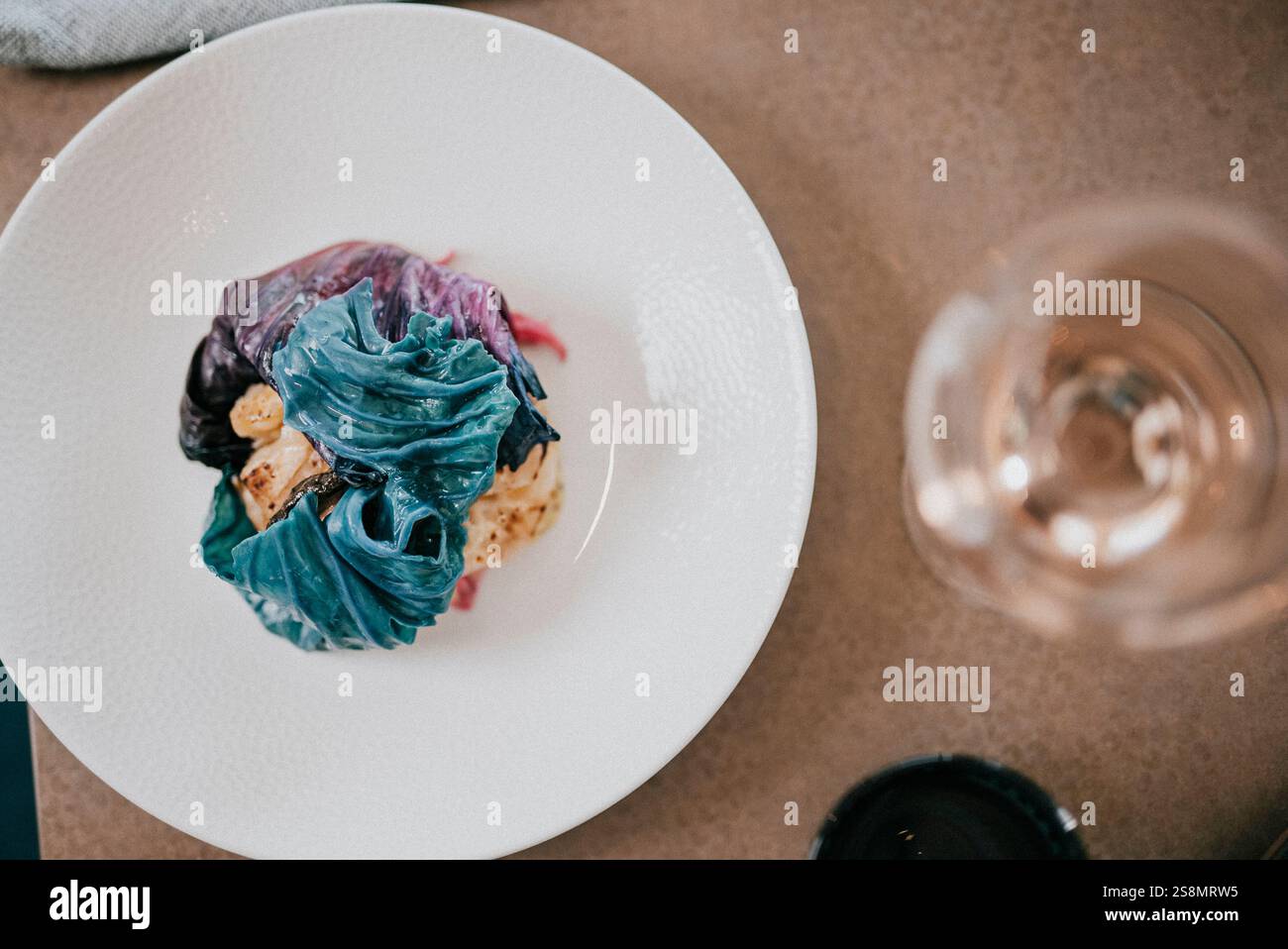 Un piatto di cibo con cibo blu si siede su un tavolo accanto a un bicchiere di vino. Il cibo sembra essere un piatto colorato con un condimento blu. Il bicchiere di vino Foto Stock