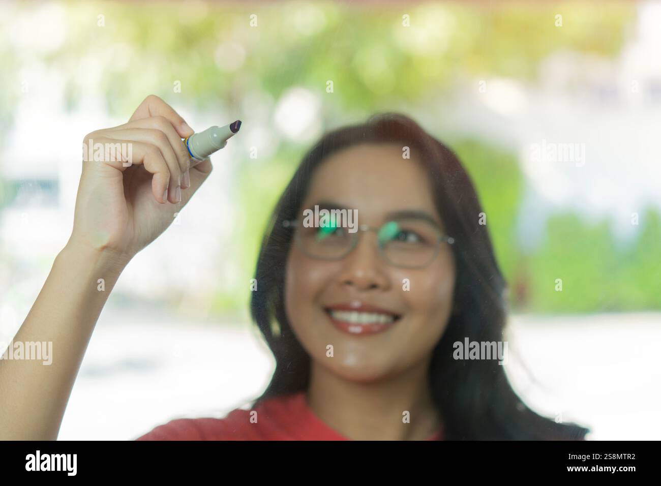 Immagine ravvicinata della giovane donna asiatica felice con una grande penna che scrive su una parete di vetro con una nuova idea. Messa a fuoco selettiva. Foto Stock