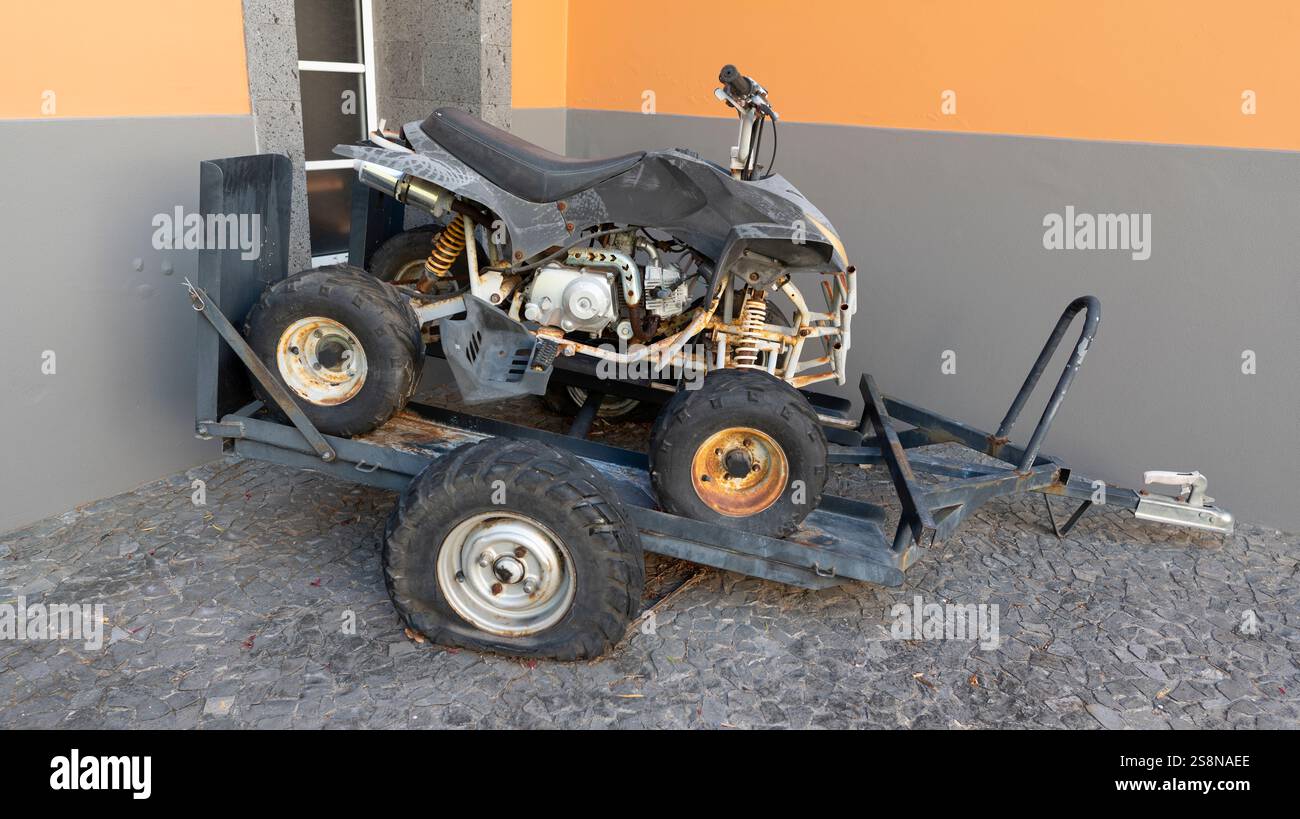 Un veicolo fuoristrada (ATV) danneggiato caricato su un rimorchio usurato, parcheggiato contro una parete colorata. Le parti rotte e lo pneumatico sgonfio suggeriscono di non riparare un Foto Stock