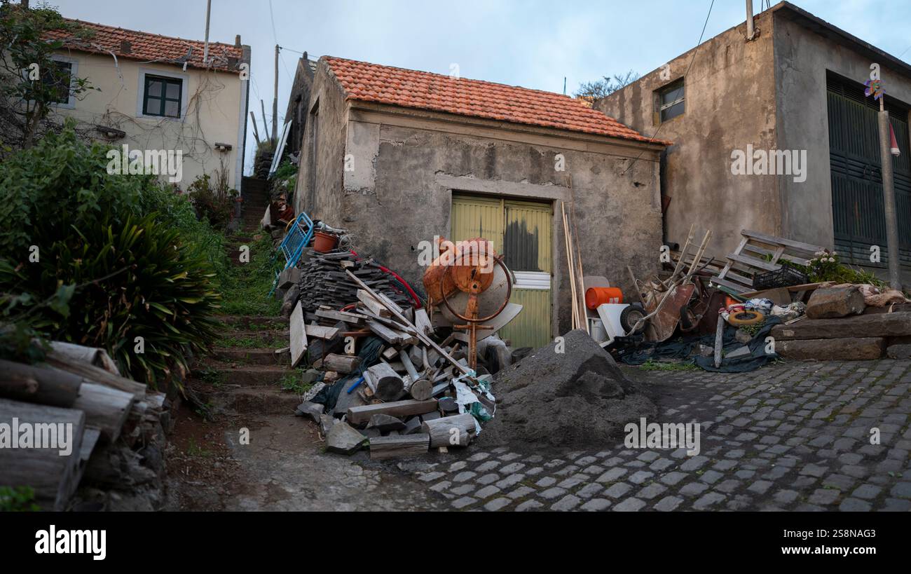 Una vecchia casa rurale con un tetto piastrellato, circondata da materiali da costruzione sparsi e oggetti scartati. Lo spazio esterno disordinato riflette un mix di Foto Stock