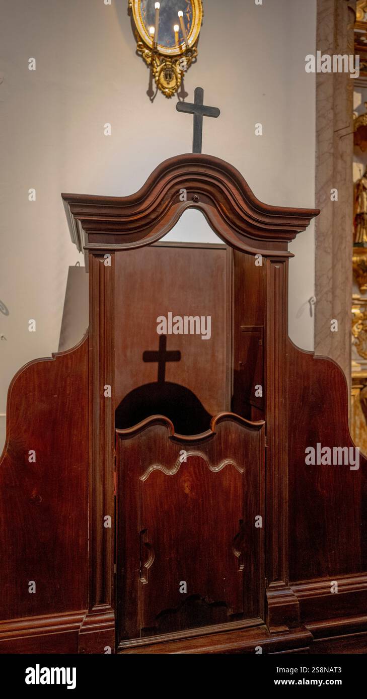 Uno stand confessionale in legno con intricati dettagli di design all'interno di una chiesa storica. Un'ombra di croce è visibile sulla cabina, illuminata dal calore Foto Stock