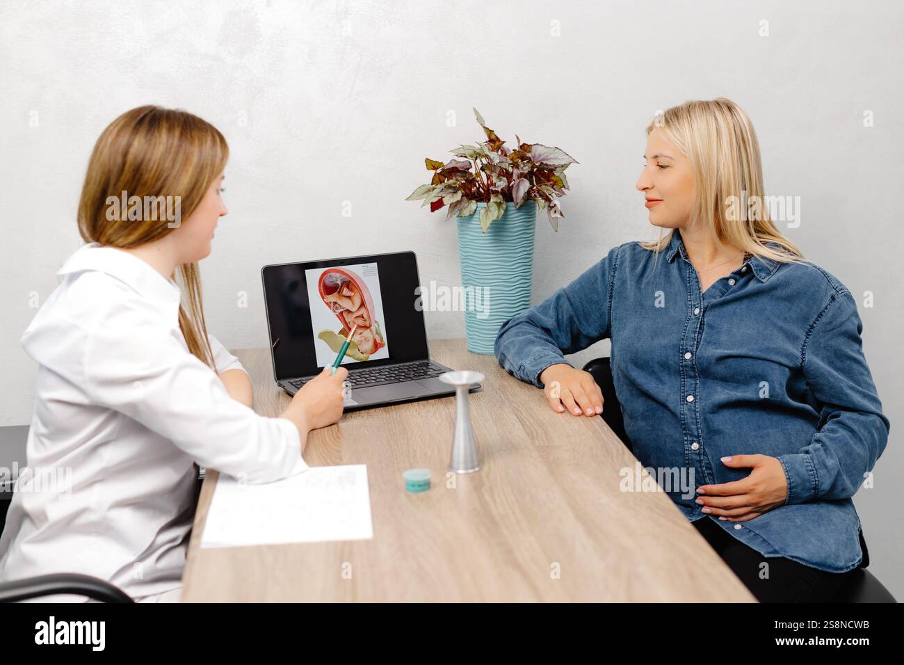 Giovane donna incinta che visita la sua ginecologa in clinica, seduta sulla sedia e parlando con il suo medico. Foto Stock