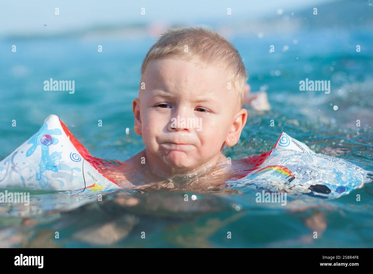 Un bambino si trova in una piscina poco profonda, indossa i galleggianti colorati e si concentra sul nuoto. L'acqua splende sotto la luce del sole, creando un allegro effetto modico Foto Stock