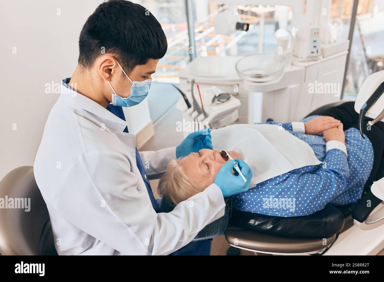 Dentista che ispeziona la bocca di una donna anziana durante le procedure dentali, le mani con i guanti garantiscono l'igiene, il paziente si rilassa sulla sedia in una clinica ben illuminata. Foto Stock