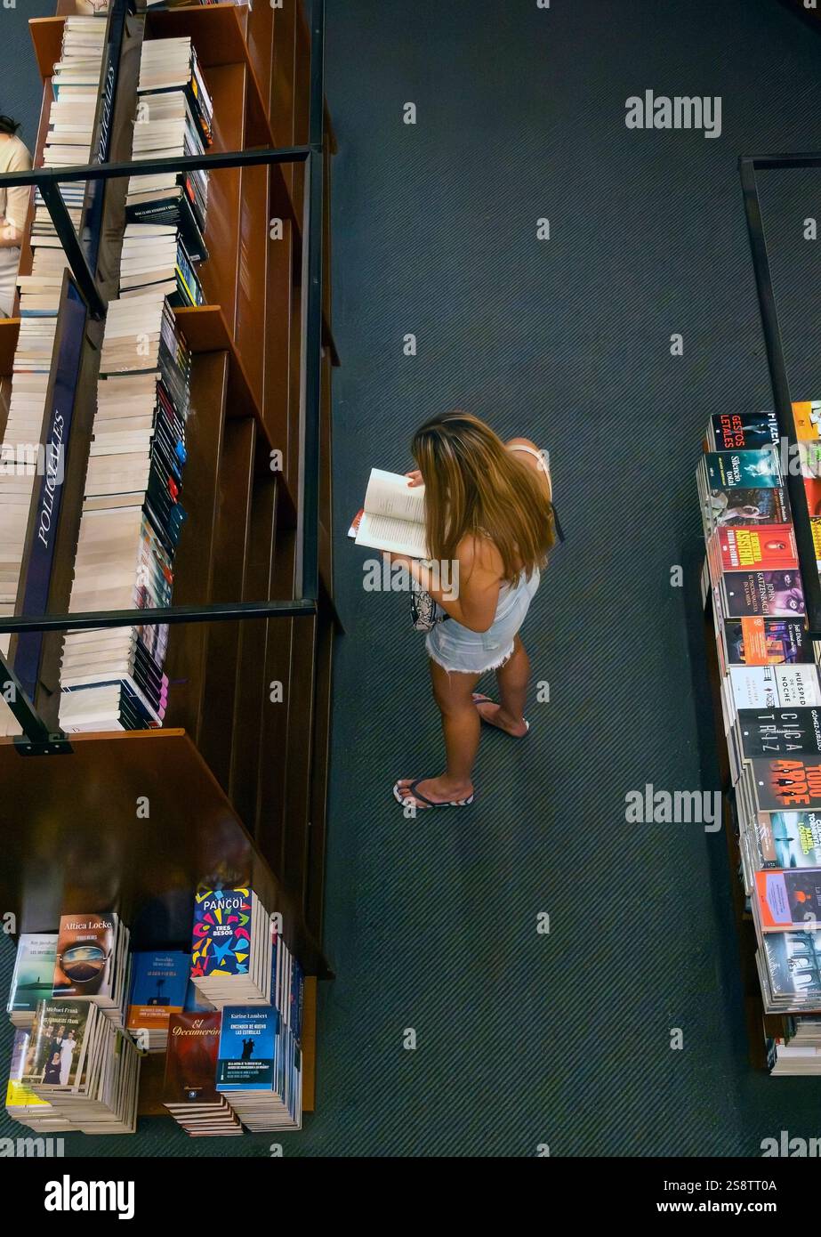 Donna bionda che indossa pantaloncini e infradito che legge un libro in libreria Foto Stock
