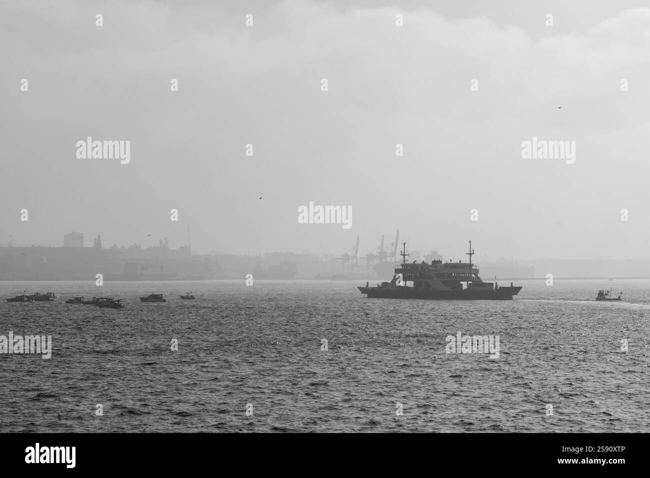 Una suggestiva vista in bianco e nero di un traghetto e di un peschereccio sul mare con uno skyline nebbioso della città sullo sfondo Foto Stock