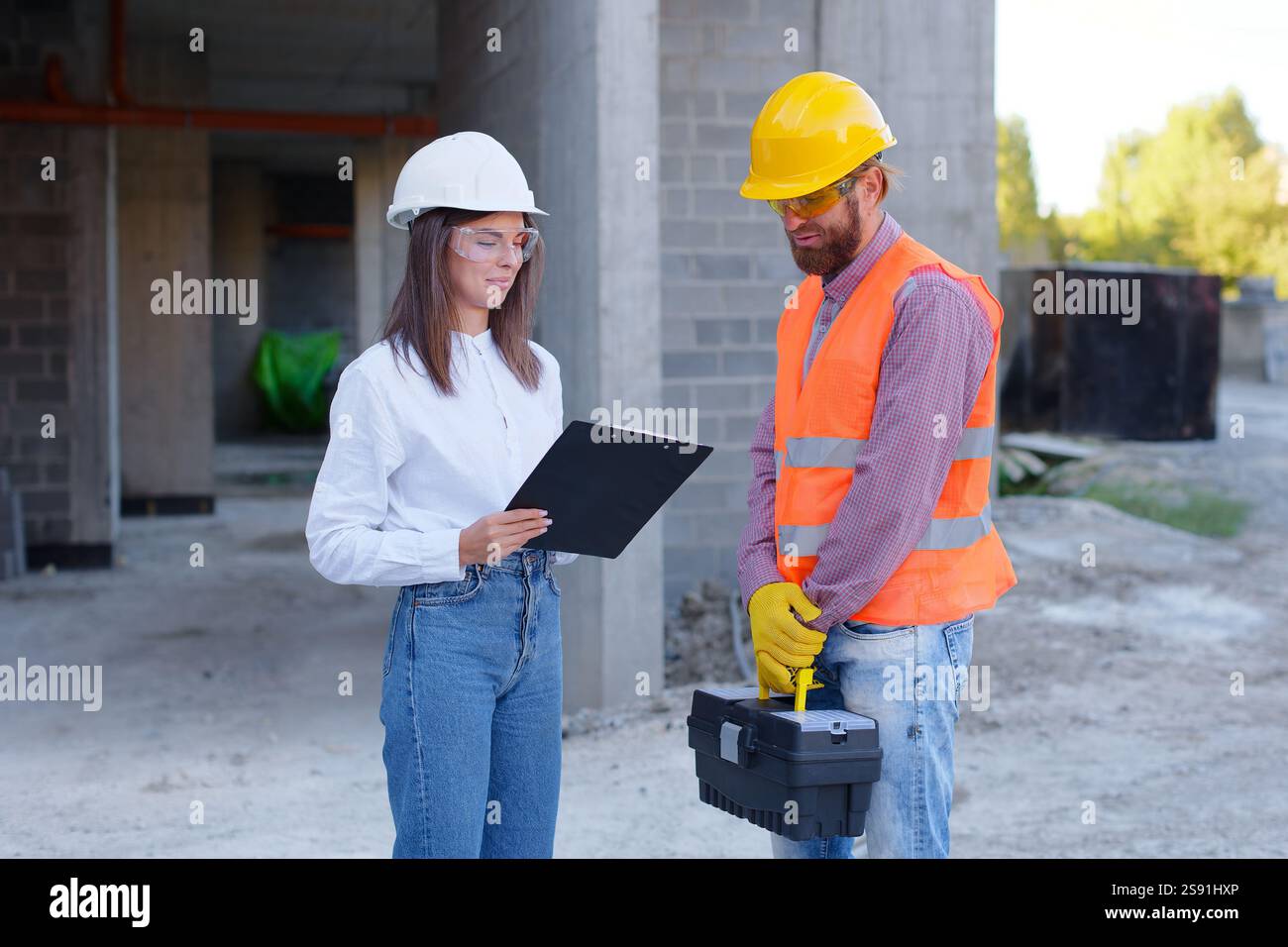Architetto che tiene appunti che discute del progetto di costruzione con il lavoratore che trasporta livella a bolla d'aria e cassetta degli attrezzi nel cantiere Foto Stock