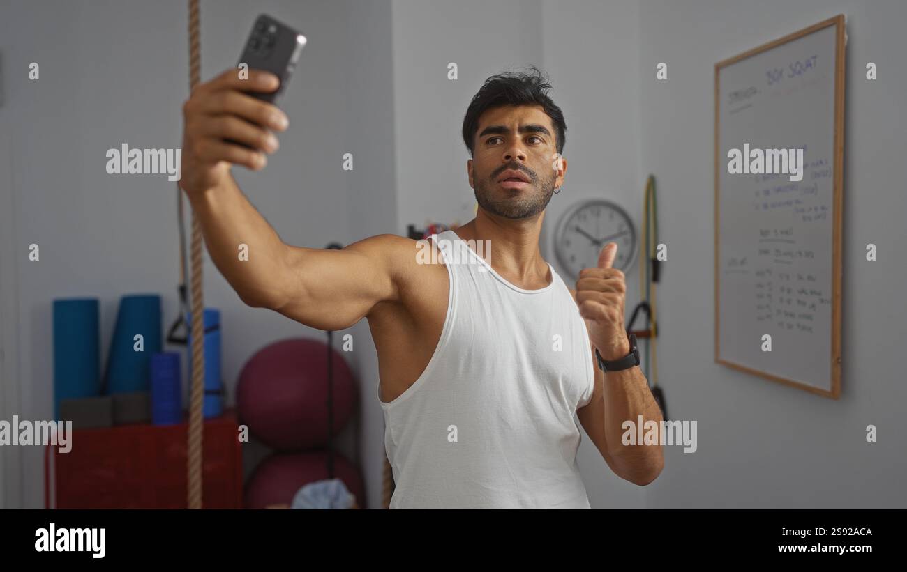 Giovane ispanico che fa un selfie in una palestra, mostra il suo fisico muscolare tenendo in mano uno smartphone e dando un pollice in su, con le attrezzature fitness Foto Stock
