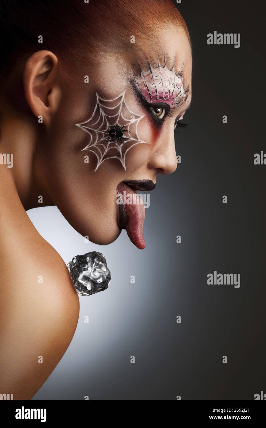 Un ritratto di una ragazza con un tatuaggio ragno di Halloween sul suo viso dipinto come una maschera spaventosa con un cubo di ghiaccio sulla spalla Foto Stock
