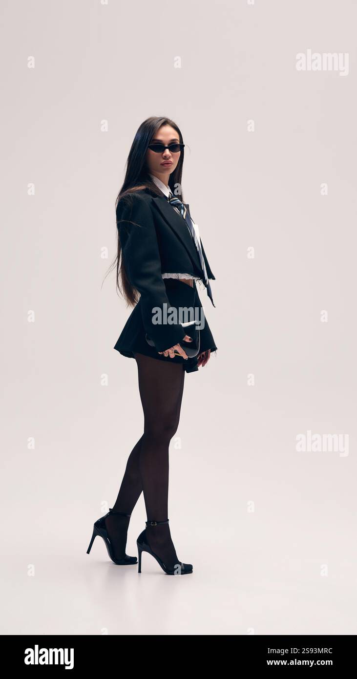 Immagine a figura intera di una bella donna che posa su una giacca corta e una camicia con cravatta e scarpe con tacco sullo sfondo dello studio Foto Stock
