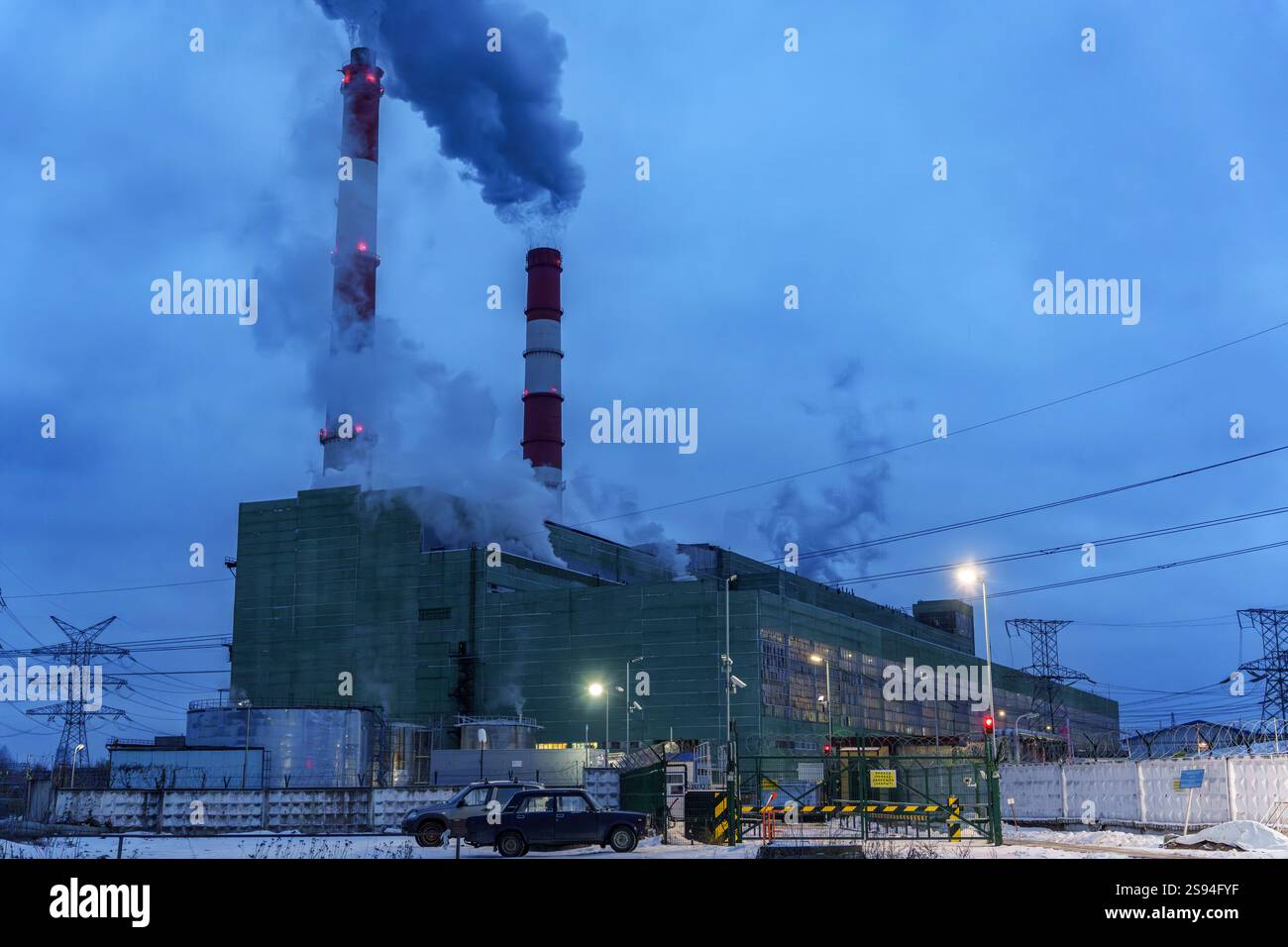 Centrale elettrica industriale con alti camini per fumo e strutture illuminate in una nevosa serata invernale, che mette in mostra la produzione di energia e l'ambiente Foto Stock
