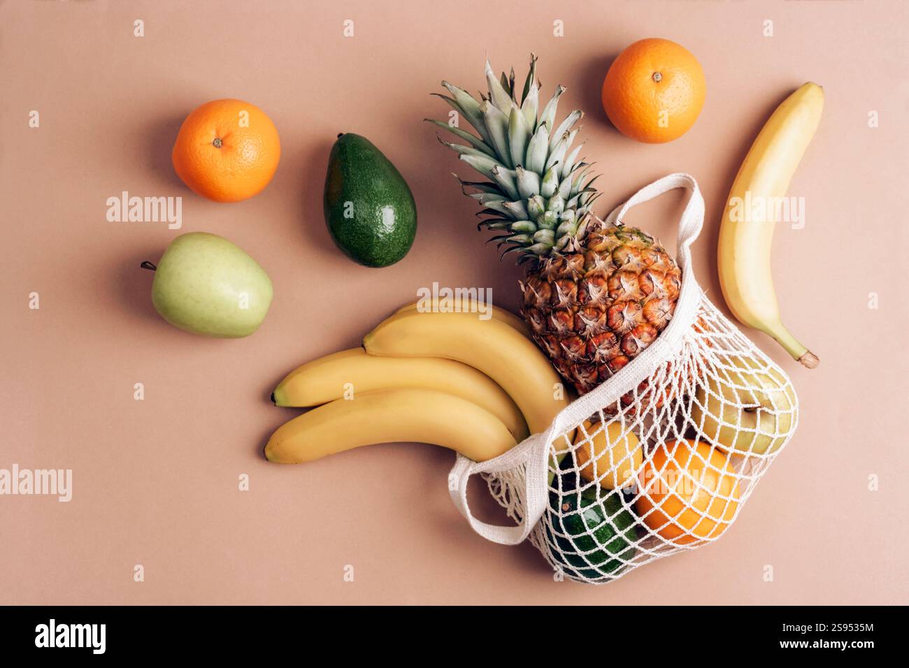 Giacimento piatto di frutta fresca: Ananas, banane, arance, avocado e mele che fuoriescono da un sacchetto in rete riutilizzabile su sfondo beige. Vista dall'alto. Foto Stock