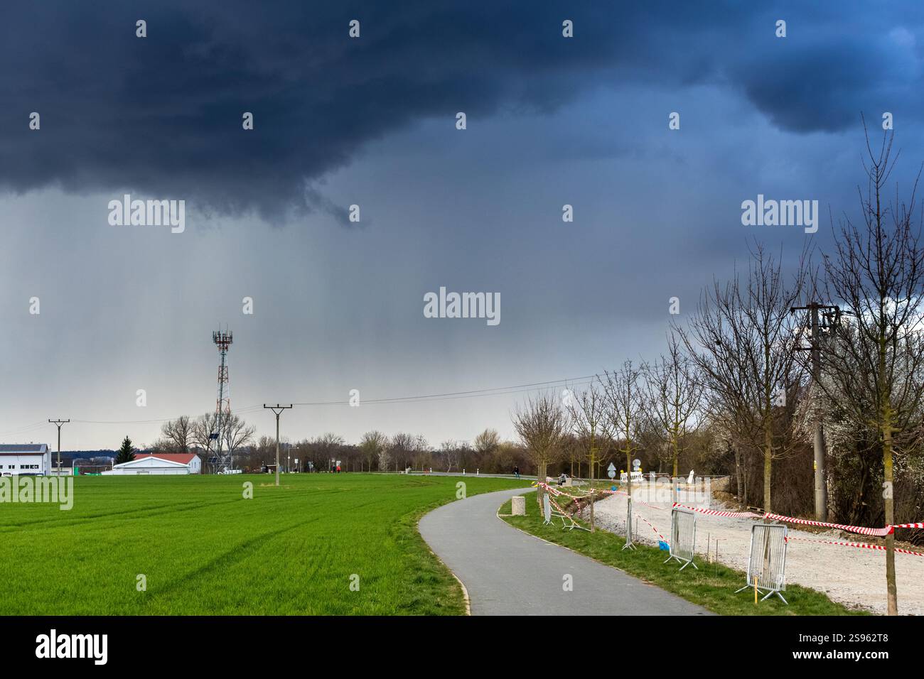 Spettacolari nuvole di tempesta su un paesaggio rurale con un campo verde, un sentiero lastricato, alberi senza foglie, una torre radio, linee elettriche, e edifici lontani. Foto Stock