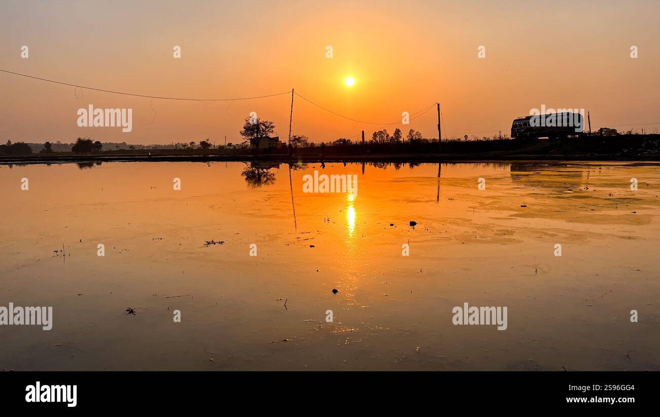 Tramonto dorato che si riflette sull'acqua con un paesaggio rurale e un autobus in movimento a Garh Mandaran, Hooghly, Bengala Occidentale, India Foto Stock