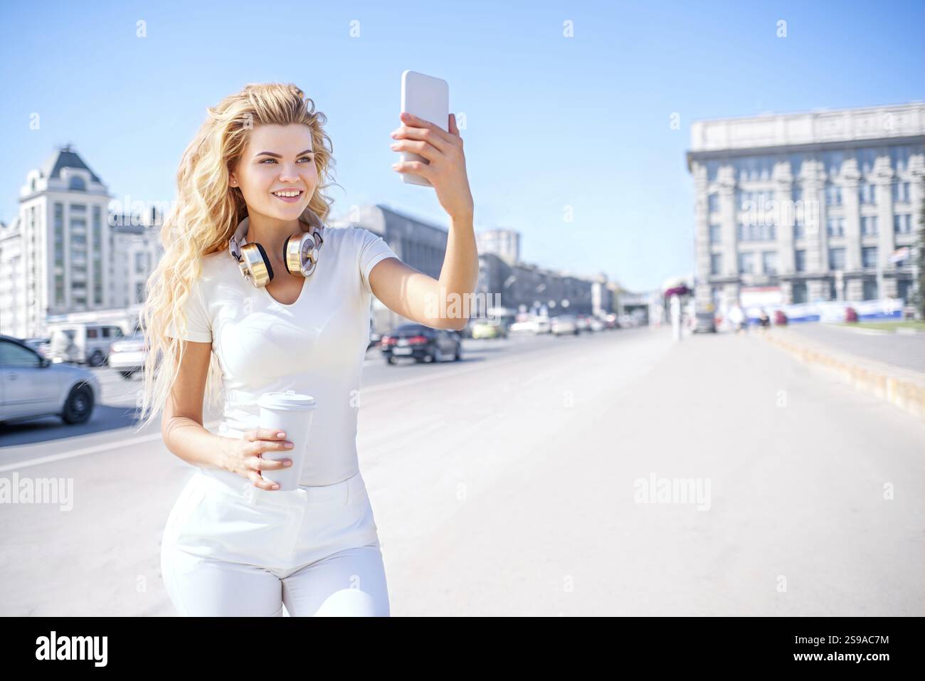 Bella giovane donna con cuffie musicali e una tazza di caffè da portare sempre con te, scattando selfie sullo sfondo urbano Foto Stock
