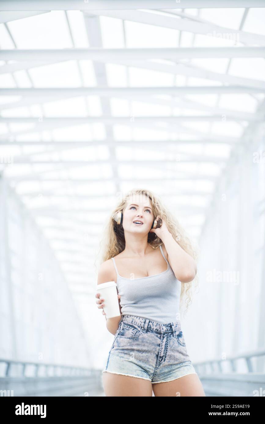 Bella giovane donna con cuffie musicali in piedi sul ponte con una tazza da caffè da asporto e in posa di fronte a uno sfondo urbano Foto Stock