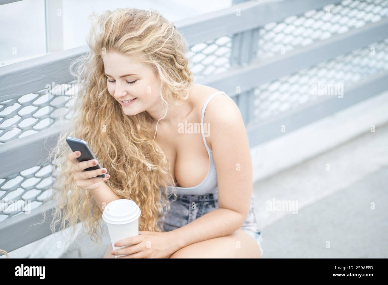 Bella giovane donna con lunghi capelli ricci e una tazza di caffè da asporto, chiacchierando e navigando al telefono sullo sfondo di un binario di metallo urbano Foto Stock