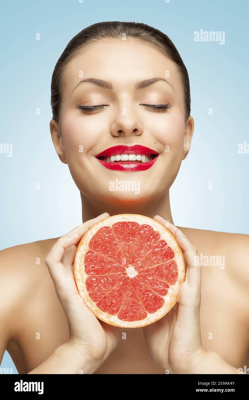 Un ritratto creativo di una bella donna sorridente che le schiaccia un pompelmo rosso nelle mani Foto Stock