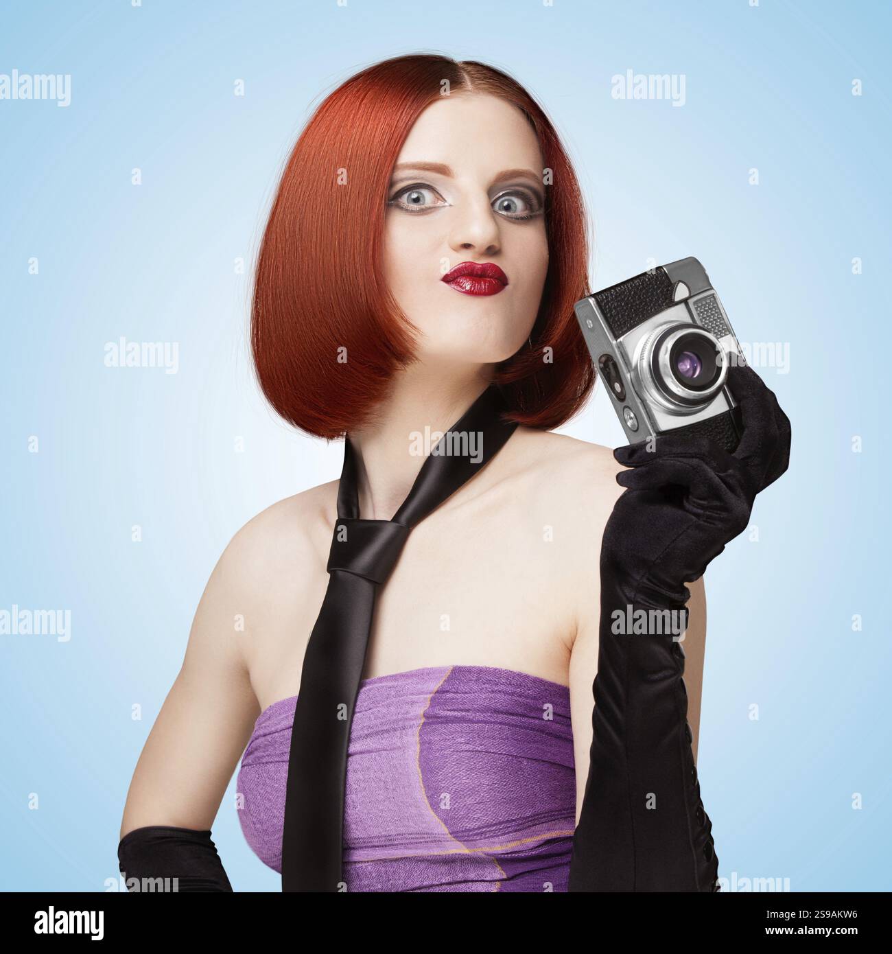 Ragazza glamour in stile retrò Vamp, con cravatta e guanti lunghi, che mostra emozioni e tiene in mano una vecchia fotocamera vintage su sfondo blu Foto Stock