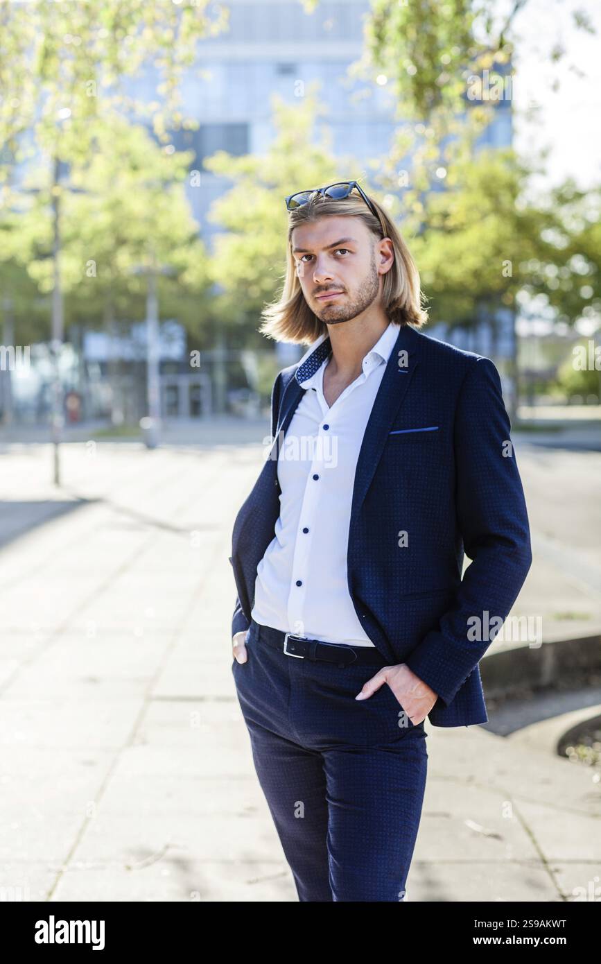 Un giovane con lunghi capelli biondi, con un elegante abito blu e una camicia bianca, posa con sicurezza all'aperto davanti a un moderno schienale architettonico Foto Stock