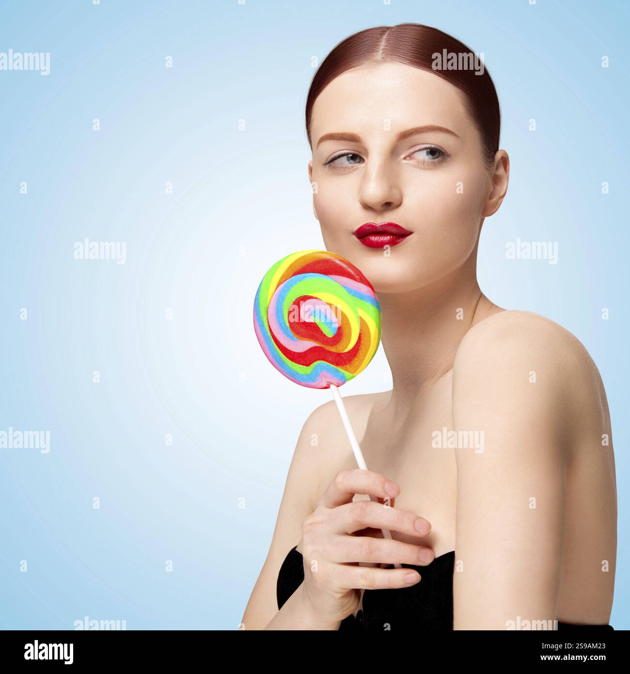Foto creativa di una bella ragazza giocosa con un lecca lecca colorato che guarda di lato e flirta su uno sfondo blu Foto Stock