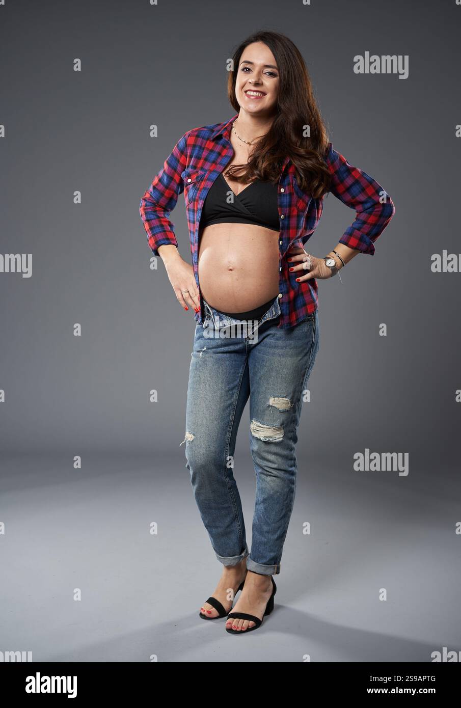 Donna incinta sicura in jeans casual e camicia aperta a quadri, sorridente e in posa con la mano sull'anca, isolata su sfondo grigio Foto Stock