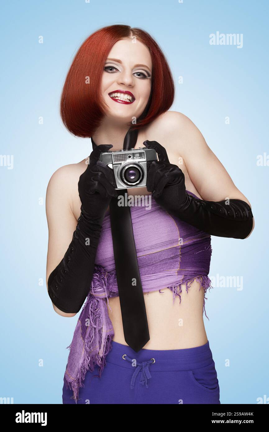 Ragazza allegra e glamour con cravatta e guanti lunghi che scatta una foto con una vecchia macchina fotografica vintage e dice formaggio su sfondo blu Foto Stock