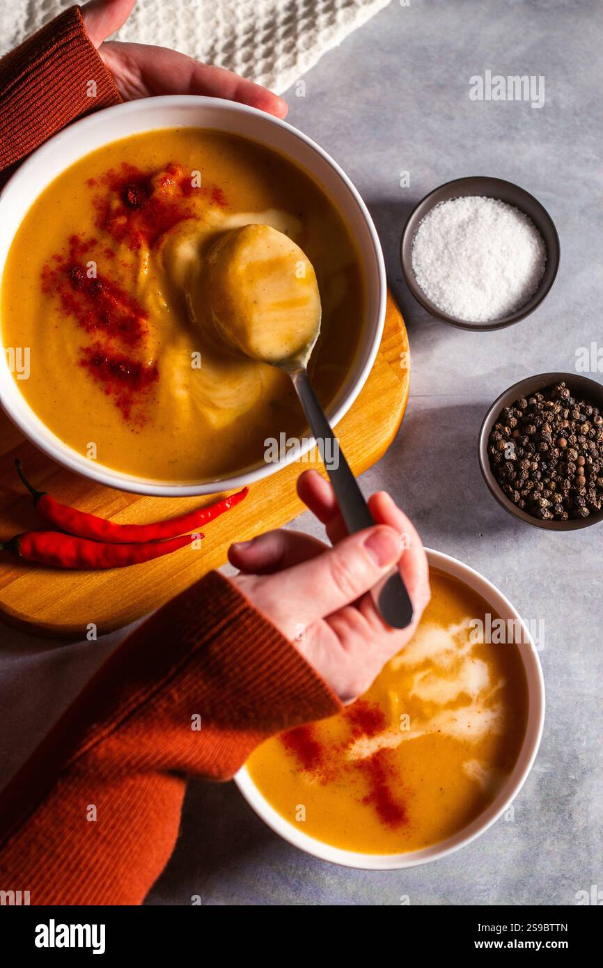 Donna che assaggia zuppa di zucca con cucchiaio. Mangiare zuppa di panna. Angolo alto. Tavolo servito con recipiente per zuppe, pepe, peperoncino, sale. Foto Stock