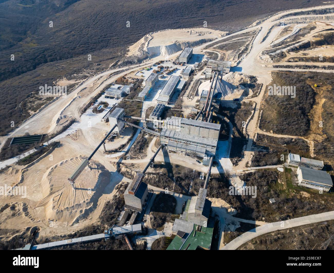 Un grande cementificio industriale circondato da colline aride e strade sterrate. I nastri trasportatori e le strutture di stoccaggio dominano il sito, mostrando attivi Foto Stock