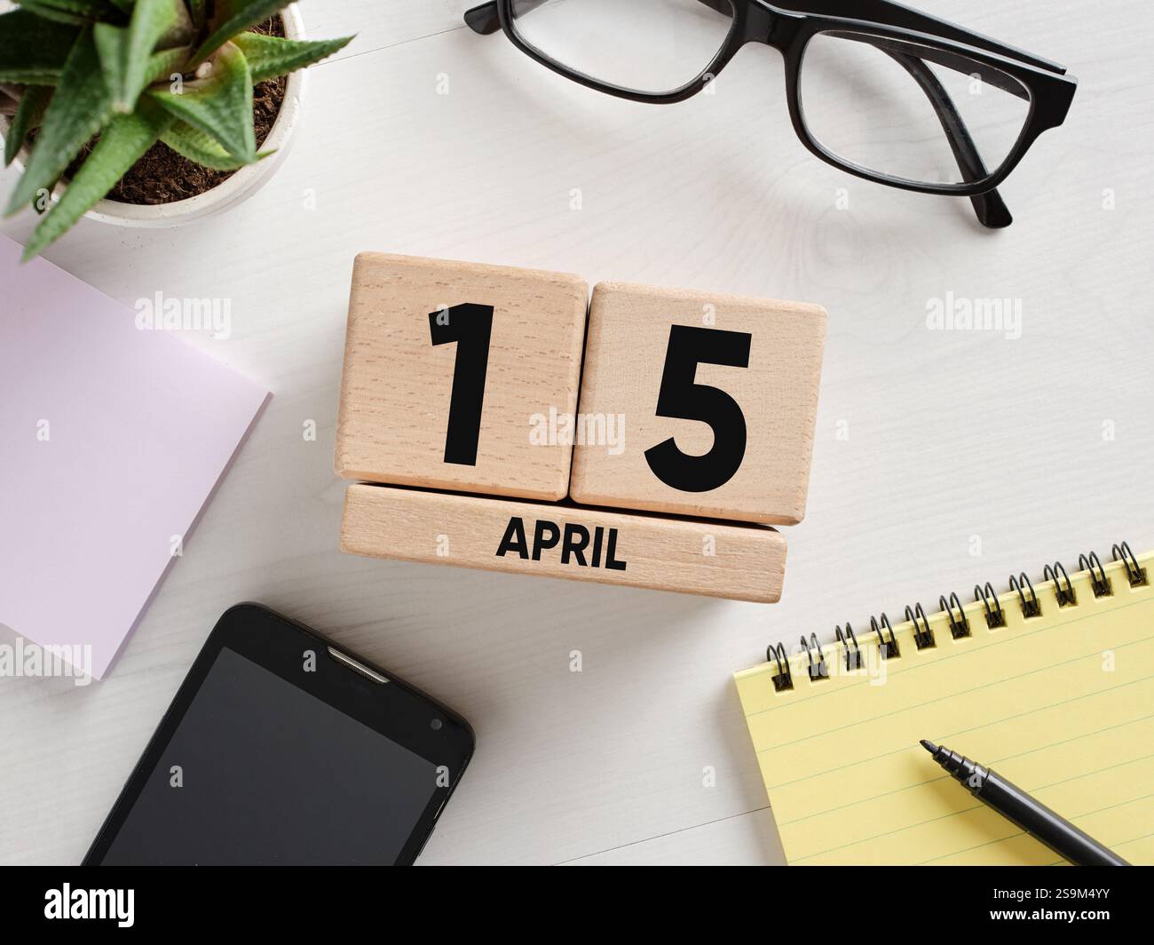 Scrivania che mostra il 15 aprile su calendario perpetuo in legno, accanto a blocco note, penna, occhiali, smartphone e impianto, ideale per promemoria fiscali Foto Stock