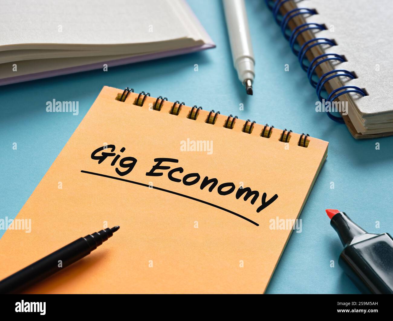 Notebook arancione con scritta Gig Economy, accompagnato da pennarelli e blocco note su una superficie blu, che rappresentano le tendenze del lavoro moderno Foto Stock