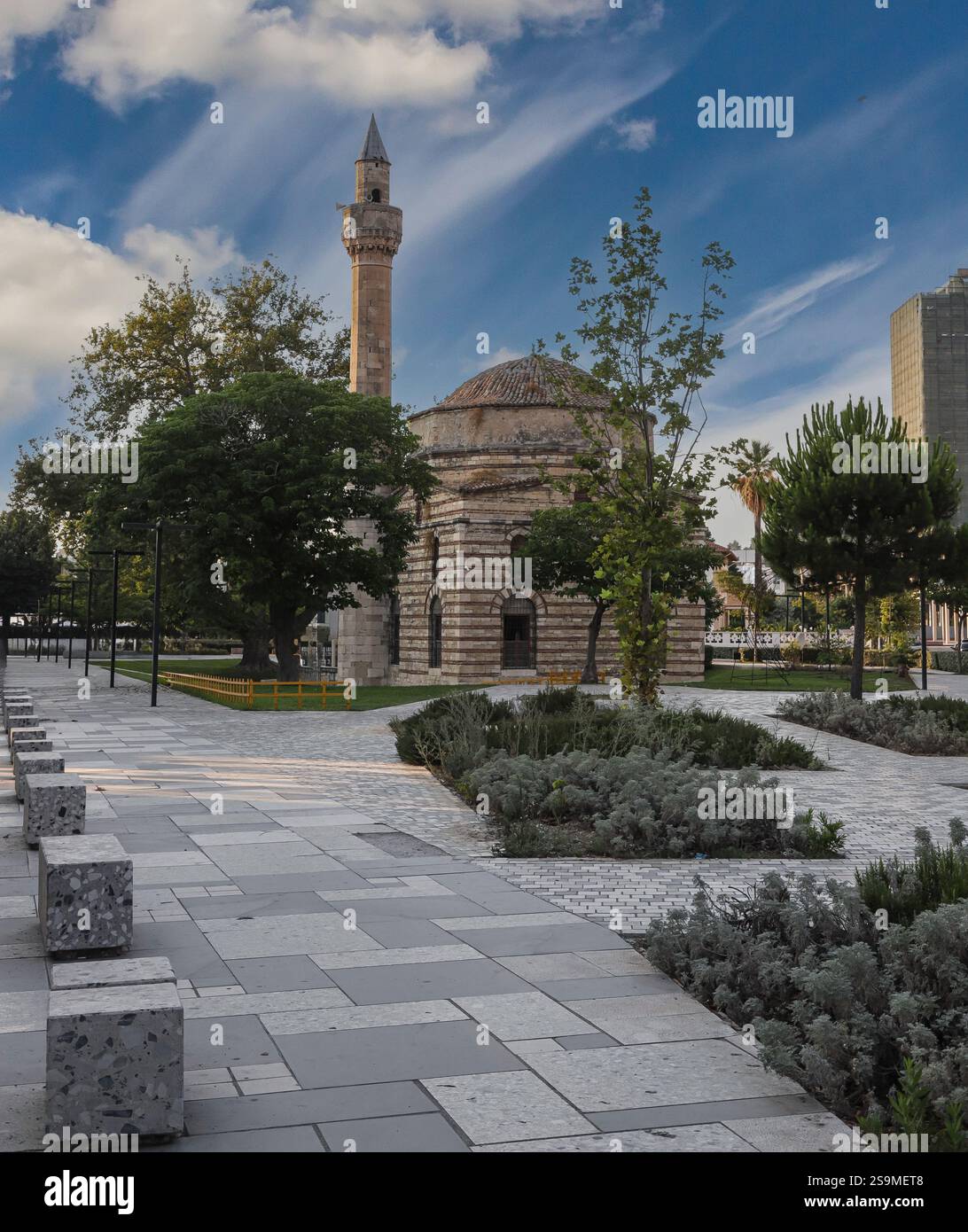 I visitatori passeggiano lungo una passerella pavimentata accanto a una moschea storica, mentre gli edifici e gli alberi moderni creano una miscela di architettura vecchia e nuova nell'ev Foto Stock