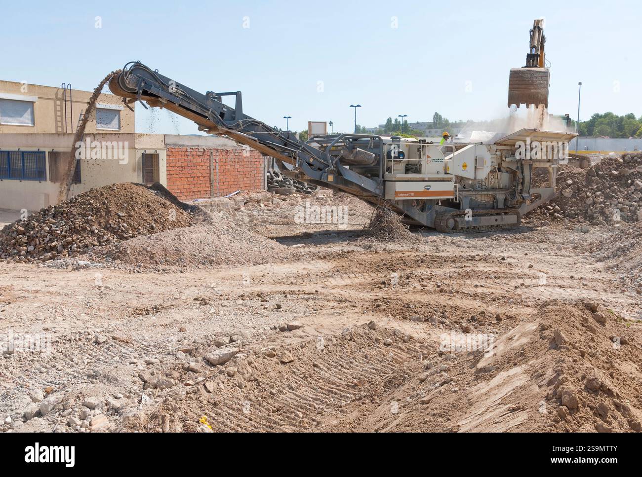 L'escavatore scarica le macerie di cemento armato nella tramoggia di un frantumatore mobile. Un lavoratore spruzza la polvere. Una cinghia magnetica separa i rottami metallici Foto Stock