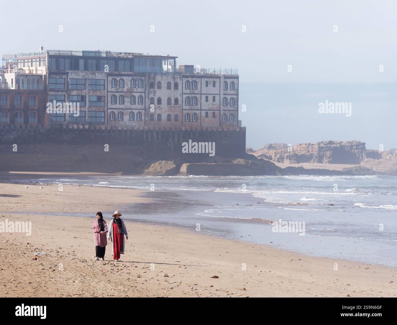 Donne musulmane che camminano su una spiaggia con la storica Medina murata alle spalle a Essaouira, in Marocco. 26 gennaio 2025 Foto Stock