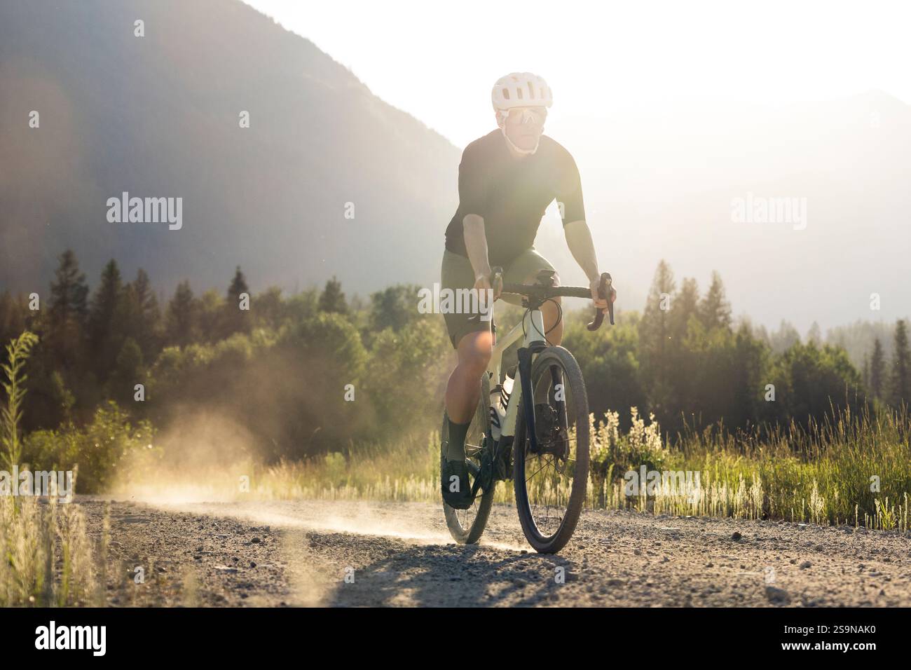 Ciclista su una strada di ghiaia in un paesaggio rurale illuminato dal sole, circondato da montagne. Foto Stock