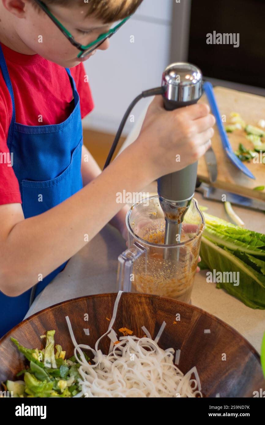 Ragazzo con grembiule blu che utilizza il frullatore a immersione per preparare l'insalata Foto Stock