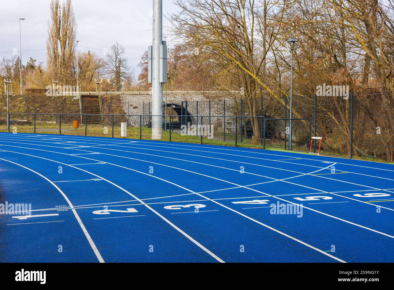 Uno stadio cittadino con piste da corsa blu e numeri di partenza bianchi. Alberi sullo sfondo. Recinzione della recinzione dello stadio. Foto Stock