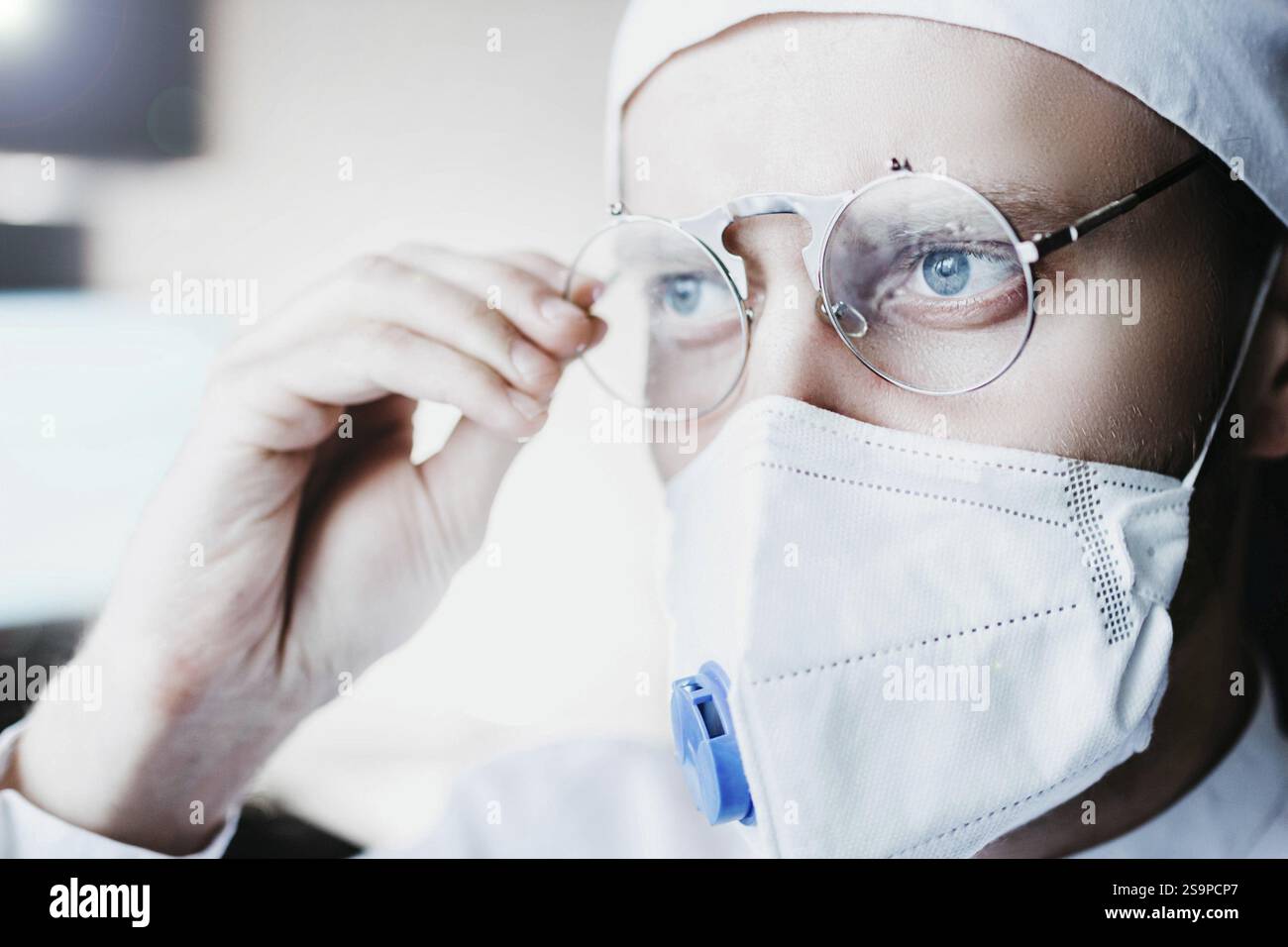 Medico che regola gli occhiali rotondi mentre indossa una maschera in ambiente clinico Foto Stock
