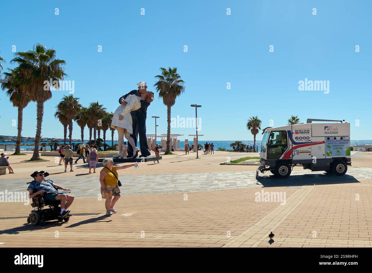 Concetto: Civitavecchia. Italia - 27 gennaio 2025: Passeggiata di Civitavecchia con statua di un marinaio che bacia un'infermiera con veicolo per la pulizia nel foregro Foto Stock