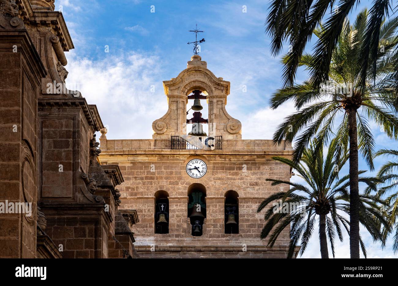 Un campanile storico con orologio, circondato da palme e un cielo blu. L'architettura presenta intricati lavori in pietra e un'importante campana. Foto Stock