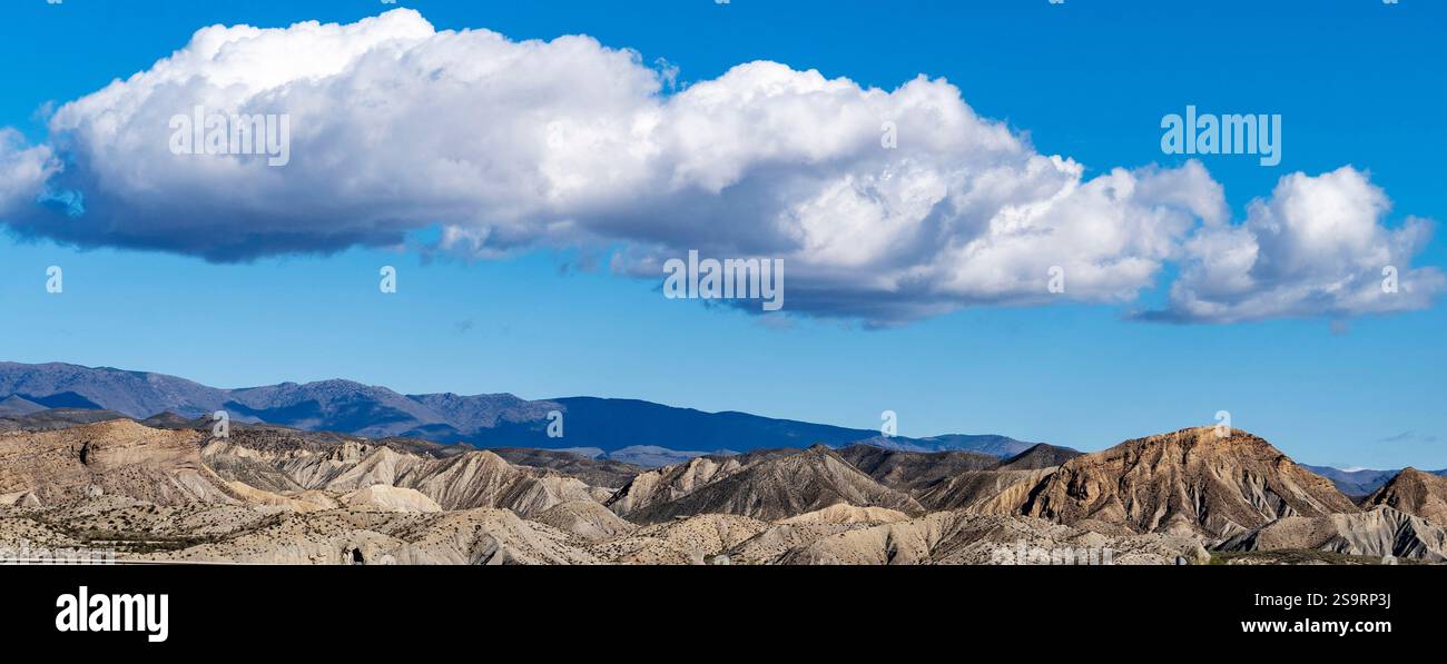 Una vista panoramica delle aspre montagne sotto un cielo azzurro luminoso con soffici nuvole bianche. Il paesaggio presenta un mix di tonalità e texture terrose, spettacolo Foto Stock