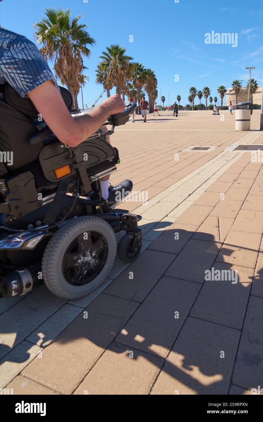 Civitavecchia. Italia - 27 gennaio 2025: Una persona su sedia a rotelle gode di una giornata di sole su un lungomare alberato di palme. La scena mostra l'accessibilità Foto Stock