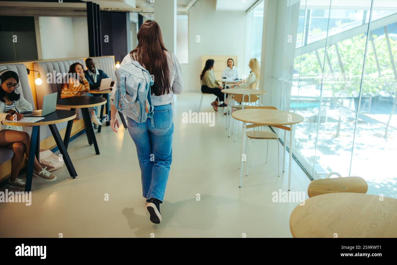 Uno studente delle scuole superiori attraversa una biblioteca cittadina, dove i colleghi sono impegnati in sessioni di studio con laptop in un ambiente luminoso e aperto. Foto Stock